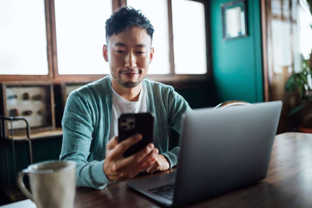 young man using Patient Self Scheduling System on smart phone sitting in front of laptop