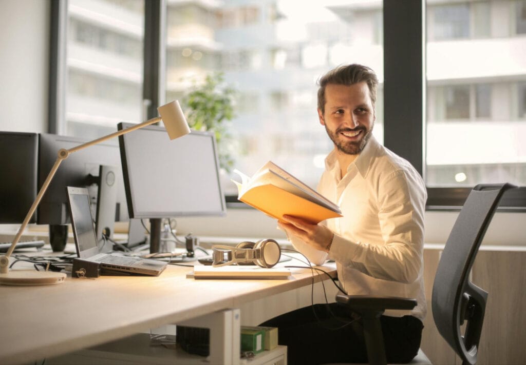 Man sitting in front of desk and computer holding a book